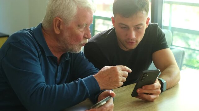 Young man assisting his elderly grandfather with a mobile phone, patiently explaining how to use a new application and navigating the touchscreen while sitting together at a wooden table at home