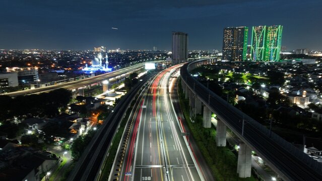 Aerial view of long exposure light trails on a highway and elevated railway tracks next to illuminated skyscrapers at night in Bekasi Barat, West Java, Indonesia.
