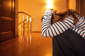 Young woman with long hair wearing striped sweater sits on wooden floor, hugging knees with head down in a softly lit indoor space © Dasha Petrenko