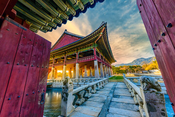 Obraz premium Beautiful view of Gyeonghoeru Pavilion in Gyeongbokgung Palace at sunset, looking through traditional red wooden gates with an ancient stone bridge in the foreground, Seoul, South Korea.