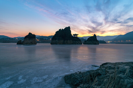 Wide angle winter sunrise over the frozen Namhan River at Dodamsambong Peaks with rocky shore foreground under a beautiful twilight sky, Danyang, South Korea.