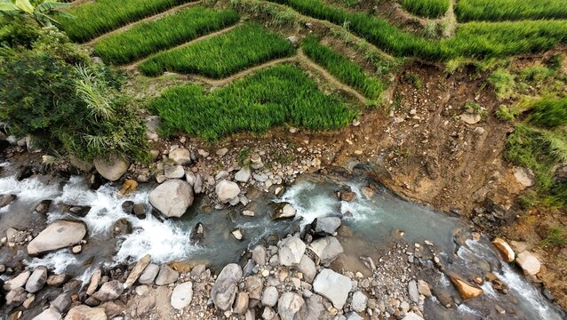 Aerial view of green terraced rice fields and a rocky river with a visible landslide area in Sukamakmur, West Java, Indonesia.