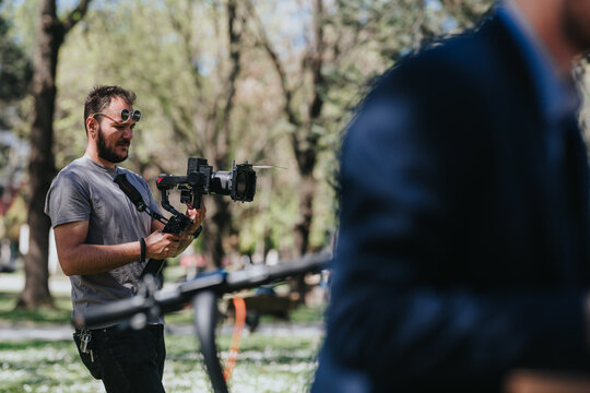 A male videographer operates a camera mounted on a gimbal while filming outdoors. The cameraman captures footage in a park setting with blurred subjects and trees in the background.