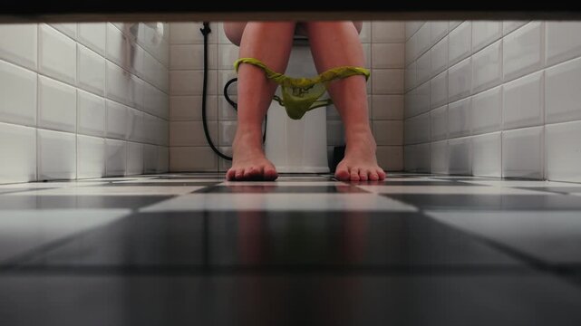 Low angle view of a woman sitting on a toilet with underwear around her ankles in a bathroom. Everyday morning routine, personal hygiene, domestic lifestyle and daily life concept. High quality 4k