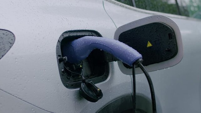 Electric vehicle charging process with blue charging cable connected to car port, rain droplets visible on the vehicle surface and charging station in the background