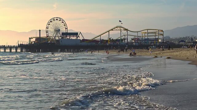 Santa Monica Pier amusement park at sunset beside ocean waves, Ferris wheel and roller coaster silhouette above a busy California beach