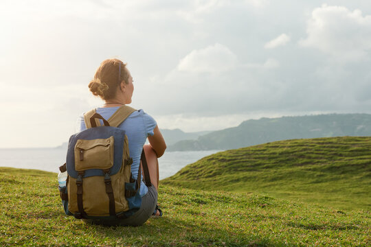 Back view of woman with canvas backpack sitting on green grass hill, contemplating mountains and sea. Solo travel concept.