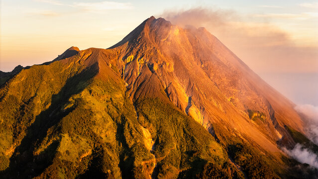 Aerial view of Mount Merapi volcano with smoke rising from the crater and golden sunlight illuminating the rocky slopes and green ridges in Indonesia.