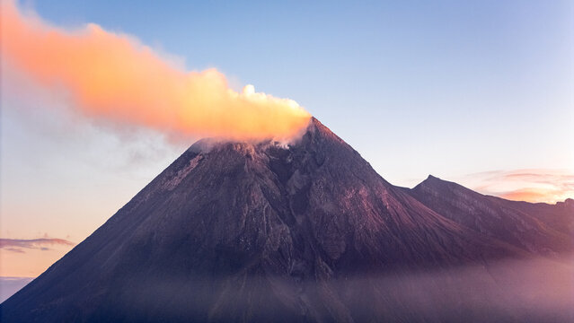 Aerial view of Mount Merapi volcano with a glowing orange smoke plume rising from the crater under a clear sky at sunrise Indonesia.