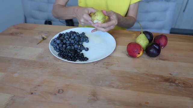 Person peeling ripe pear above plate with blueberry and currant berries in kitchen video Full HD