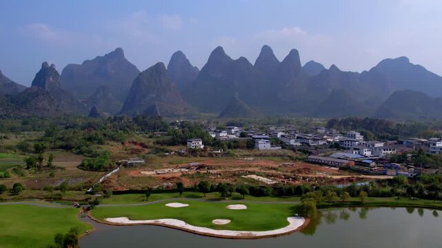 A scenic view of a karst mountain range, a village, and a golf course with a lake on a clear day.