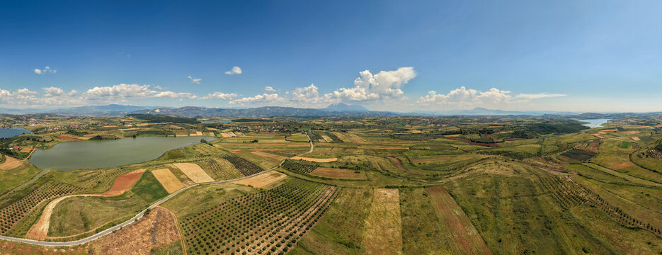 Aerial view of the rolling hills and patchwork agricultural fields surrounding a calm lake under a clear blue sky with distant mountains Belesh, Qarku i Elbasanit, Albania.