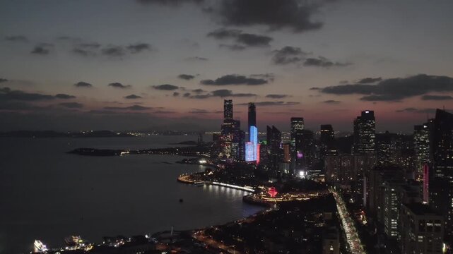 A dusk-to-night aerial view of a coastal urban skyline with illuminated skyscrapers and calm water reflecting city lights.