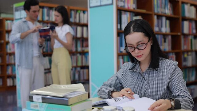 A focused young Asian female college student wearing glasses sits at a desk in a bright university library, carefully reading a textbook with a stack of books and headphones nearby.