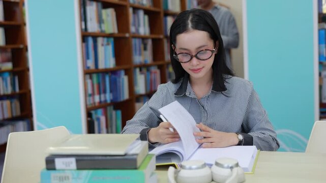 A focused young Asian female college student wearing glasses sits at a desk in a bright university library, carefully reading a textbook with a stack of books and headphones nearby.