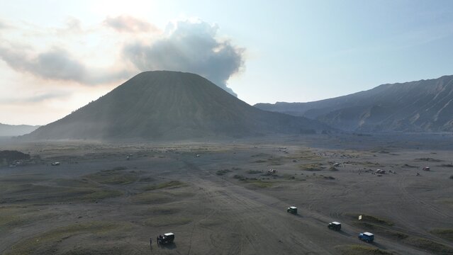 Aerial view of Mount Bromo volcano with smoke rising from the crater and off-road vehicles driving across the sandy plain in Tosari, East Java, Indonesia.