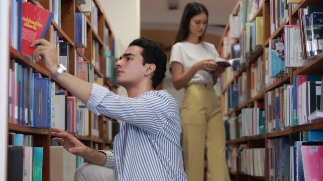 A dedicated young male college student kneels in a library aisle to read a book he found, while a female classmate searches the tall bookshelves in the blurred background.
