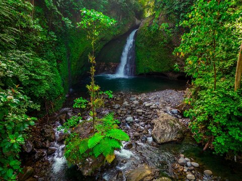 Aerial view of Palak Siring Kemumu waterfall cascading into a natural pool surrounded by dense tropical rainforest and rocky stream in Kecamatan Arma Jaya, Bengkulu, Indonesia.
