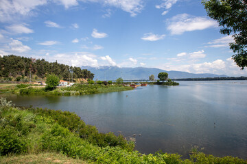Fototapeta premium Wide view of a calm lake with distant mountains, blue sky and green shoreline in Greece.