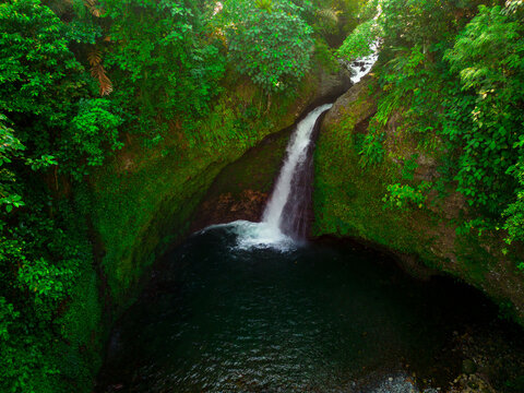 Aerial view of the Palak Siring Kemumu waterfall cascading into a dark pool surrounded by lush tropical vegetation and mossy cliffs in Kecamatan Arma Jaya, Bengkulu, Indonesia.