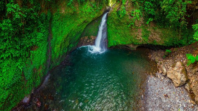 Aerial view of Palak Siring Waterfall cascading into a turquoise pool surrounded by lush green mossy cliffs in Kecamatan Arma Jaya, Bengkulu, Indonesia.