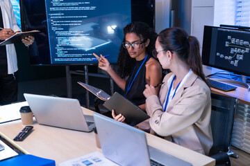 Naklejka na ściany i meble Focused female software engineer discuss project script via laptop. Diverse tech startup coder woman use tablet to check computer program code and web database development in office meeting