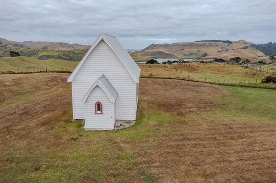 Aerial view of the historic Awhitu Central Church with its white wooden siding set against rolling hills and a distant lake under an overcast sky in Awhitu, Auckland, New Zealand.