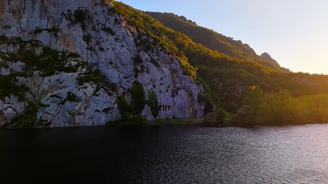 Smooth slow-motion 4K 60fps drone movement from left to right over a river at sunset, showcasing ancient royal tombs carved into the cliffs. Cinematic golden hour lighting with a calm and historic lan
