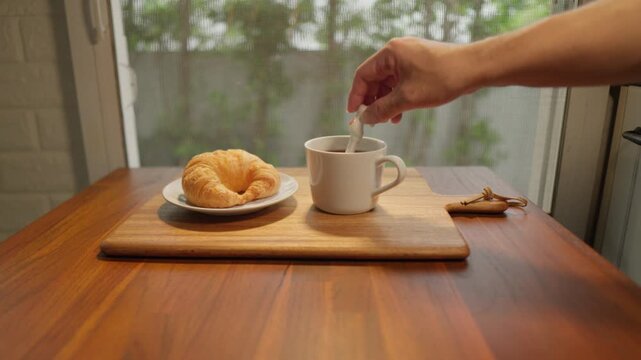 A dolly camera in slow motion captures a wooden tray with a cup of freshly brewed coffee and a croissant. A spoon is then used to stir the coffee, creating a delicious breakfast amidst the soft mornin