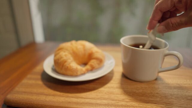 A dolly-out slow-motion camera captures a wooden tray with a cup of freshly brewed coffee and a croissant. A spoon is then used to stir the coffee, creating a delicious breakfast amidst the soft morni