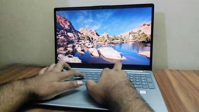 Hands typing on silver laptop keyboard during day indoors with desert scenery screen saver.