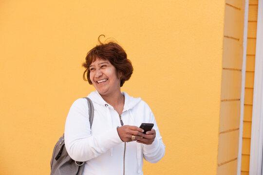 A senior woman stands against a yellow wall holding a cell phone