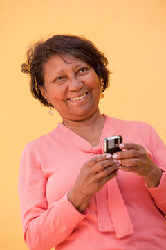 A senior woman stands against a yellow wall holding a cell phone
