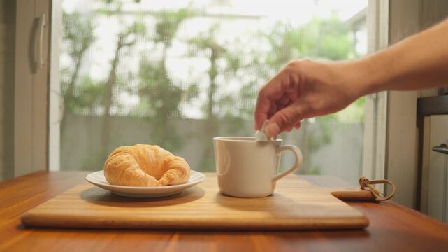 The dolly-in camera features a wooden tray with a cup of freshly brewed coffee and a croissant. A spoon is then used to stir the coffee, creating a delicious breakfast amidst the soft morning sunlight