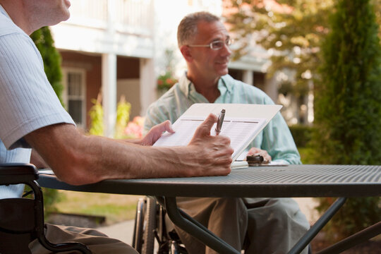 Man with Friedreich's Ataxia holding a pen with degenerated hands sitting with a man with spinal