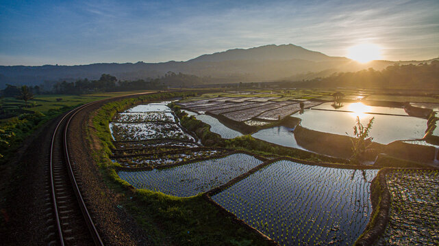 Aerial view of a curved railway track running alongside flooded rice terraces with mountains in the background during a golden sunrise Indonesia.