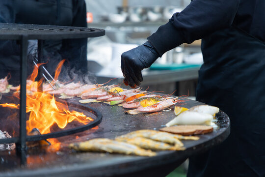 Unrecognizable chef wearing black gloves and apron grilling fresh prawns, fish, and squid on a large outdoor fire pit grill