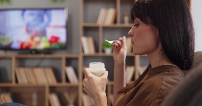 Woman eating yogurt at home, watching television. Cozy living room scene shows a routine with a yogurt cup and spoon, highlighting healthy breakfast nutrition. Clear concept of healthy lifestyle.