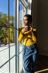 African American woman standing by multi-pane window at home holding ceramic mug in yellow top © wavebreak3