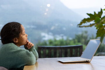 Fototapeta premium Female child in green sweater gazing through glass wall at valley, laptop on table, copy space