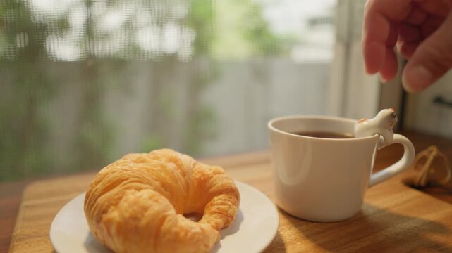 The camera moves from left to right. A wooden tray with a cup of freshly brewed coffee and a croissant is placed down. A spoon is then used to stir the coffee, creating a delicious and refreshing brea