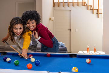 Fototapeta premium Diverse female friends holding cue stick, lining up shot on blue pool table in living room