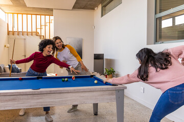 Obraz premium Three female friends playing pool on blue-felt pool table at home, using wooden cues now