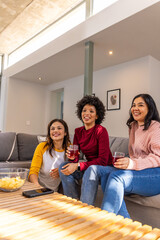 Fototapeta premium Diverse female friends sitting on gray sectional sofa in modern living room holding clear glasses
