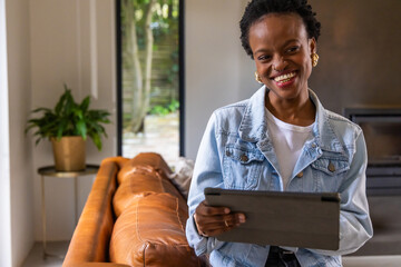 Obraz premium African American adult woman at home wearing denim jacket holding tablet near sofa, copy space