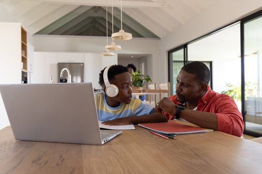 African American father and teen son using laptop while pointing at watch at wooden kitchen table