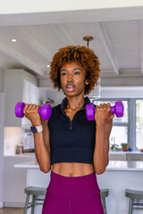 African American woman lifting purple dumbbells in kitchen in crop top and leggings with smartwatch