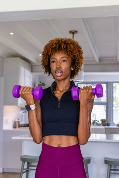 African American woman lifting purple dumbbells in kitchen in crop top and leggings with smartwatch