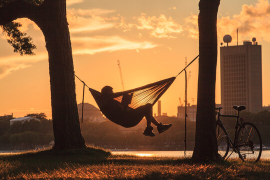 A young man sits reading in a hammock between two trees at the water's edge at sunset with a