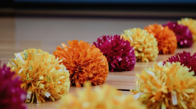 Vibrant colorful cheerleading pom poms rest on a polished wooden gym floor in a row. These fluffy shiny objects are ready for a school pep rally or athletic event.